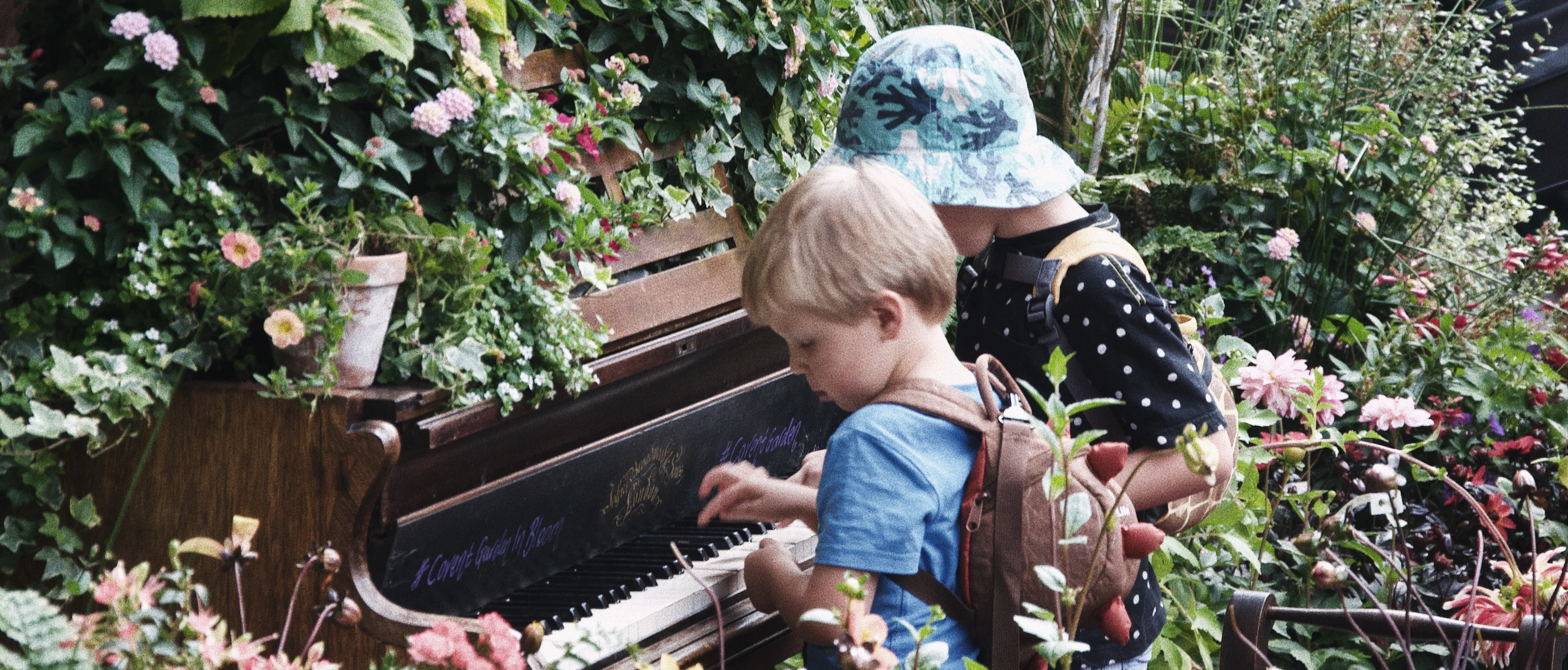 Two children at an upright piano surrounded by garden flowers.