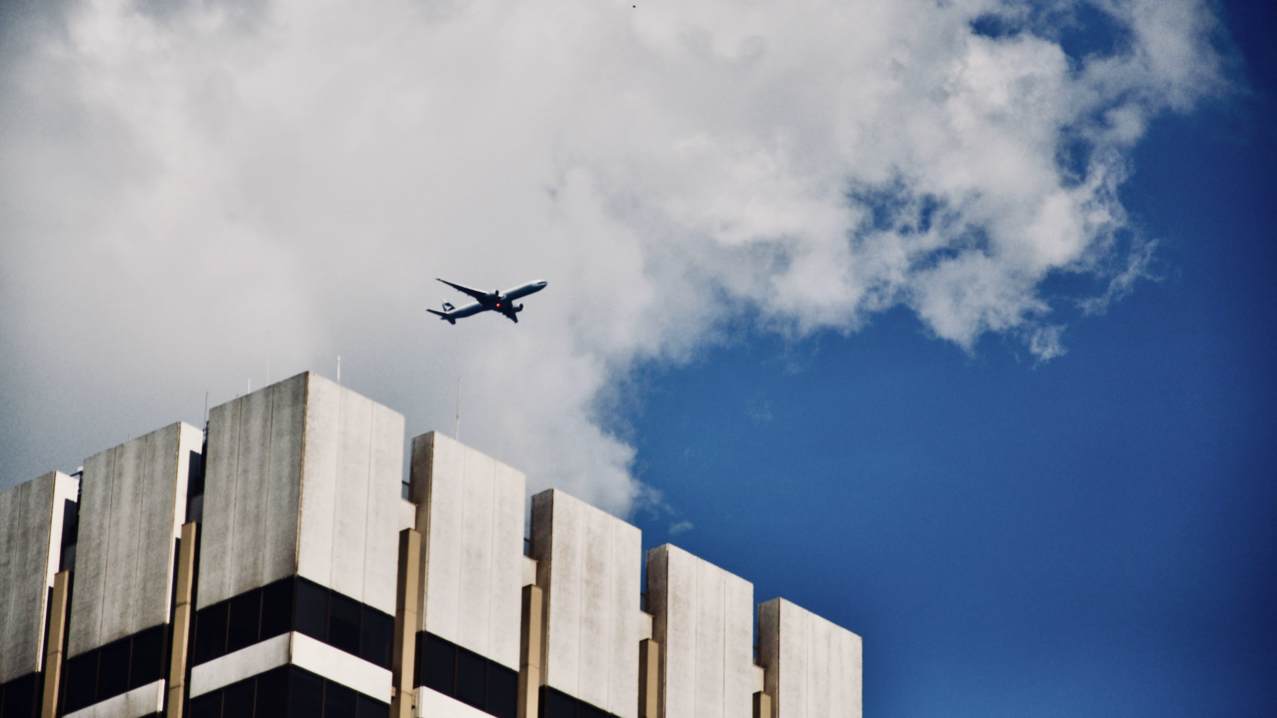 An airplane passing over a brutalist concrete building against blue sky.