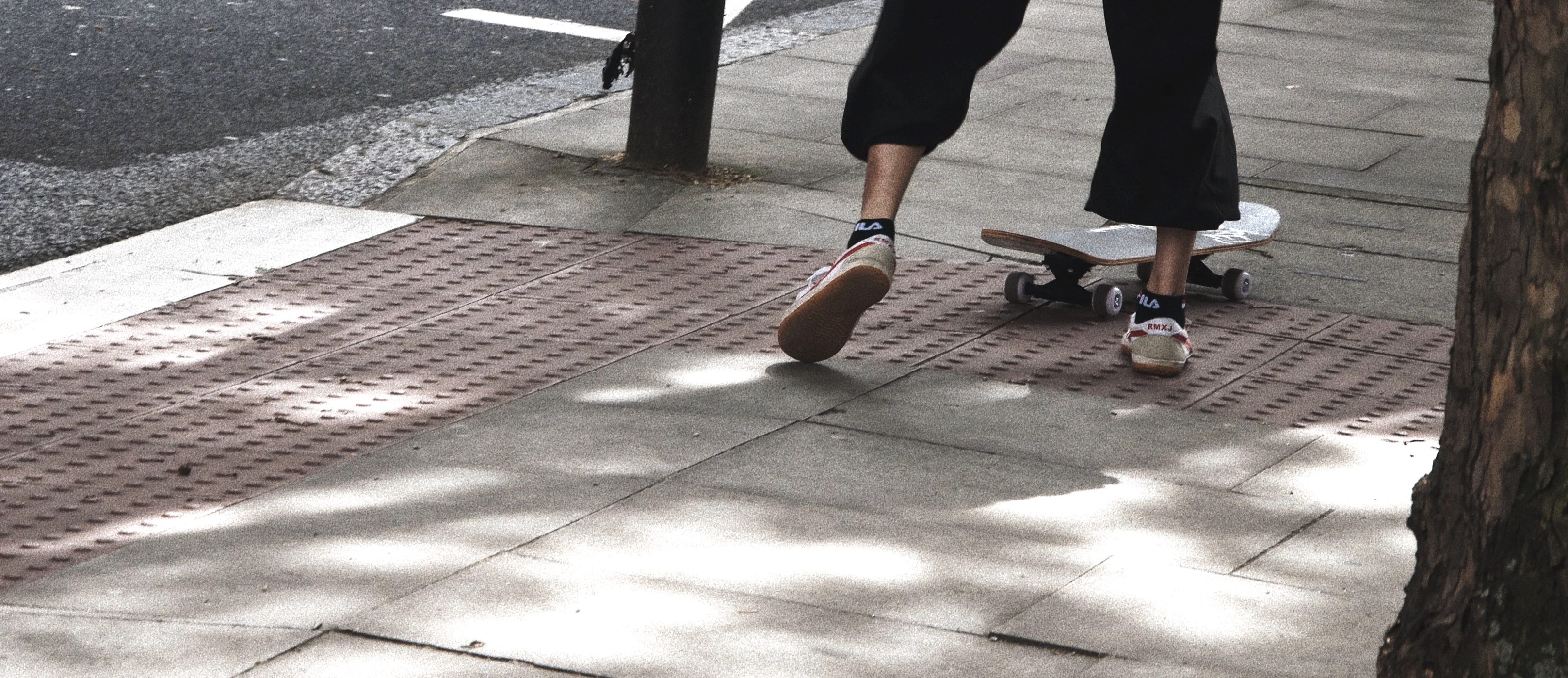 Feet and a skateboard in dappled tree shadow on a London pavement.