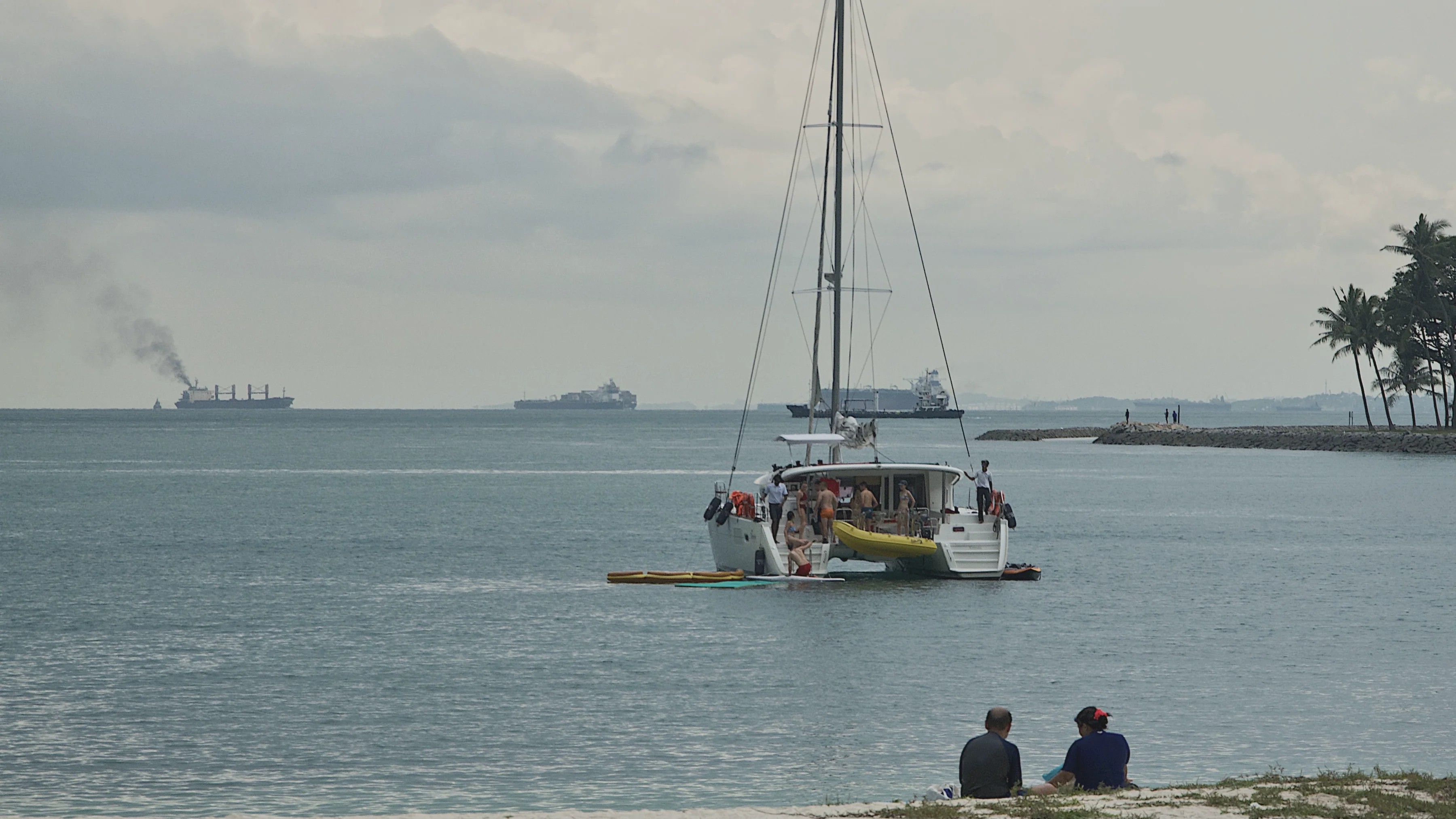A sailboat near shore with cargo ships on the horizon in Singapore.