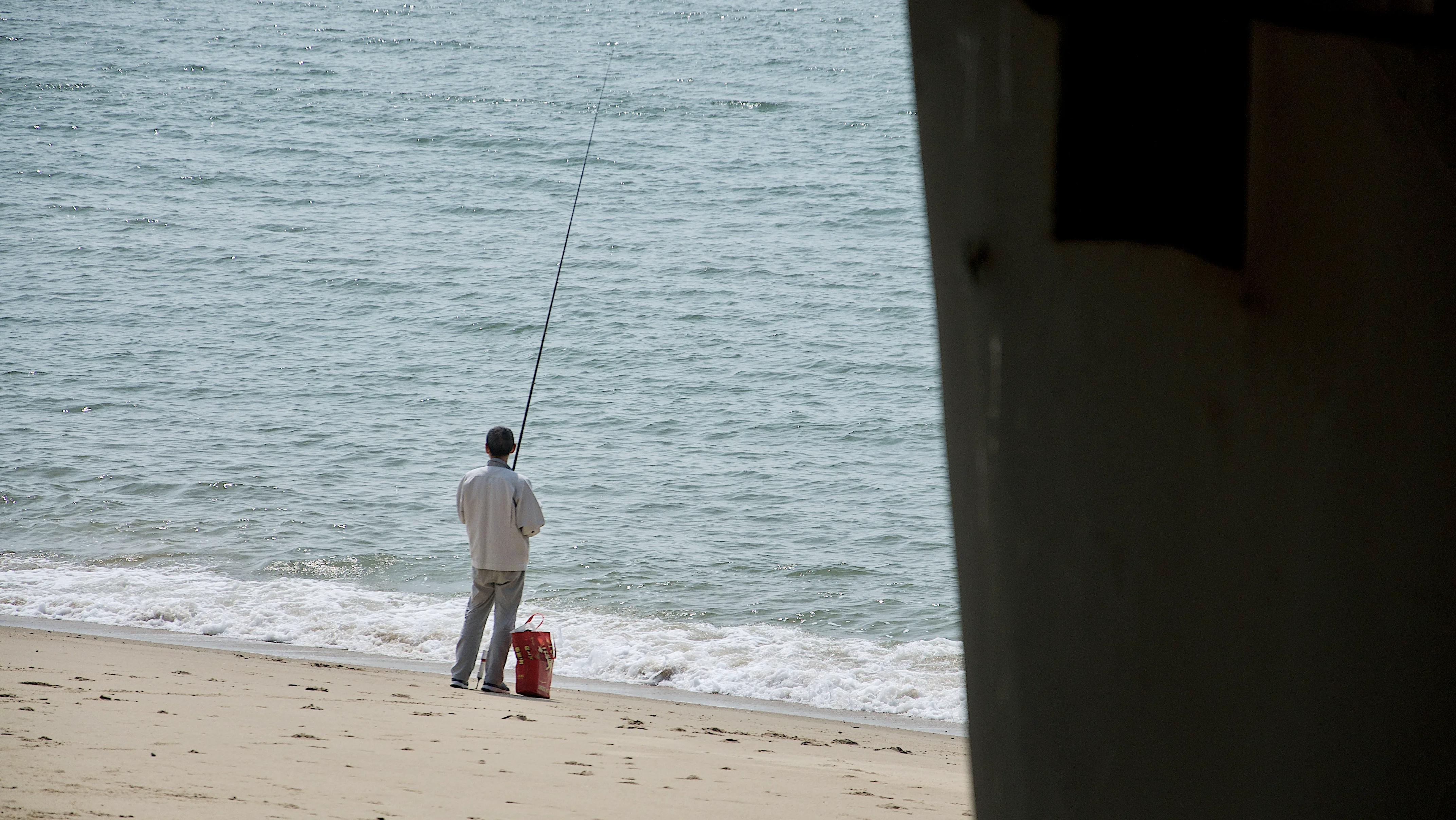 A person fishing on a Xiamen beach beneath a pier.