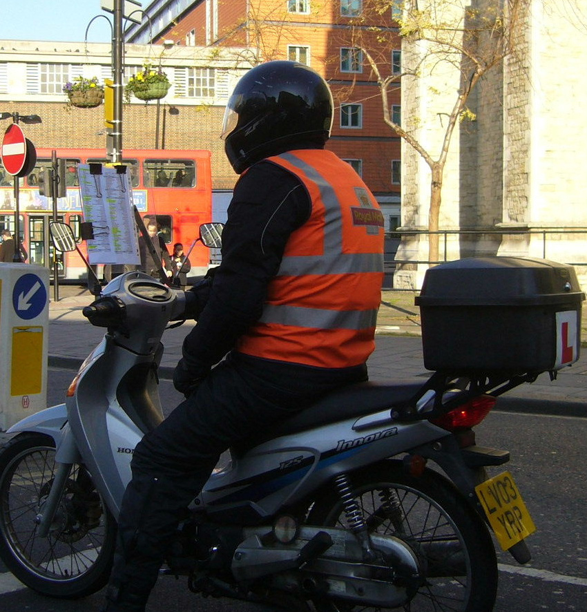 A Knowledge boy on a motor scooter at traffic lights, studying street routes on a clipboard